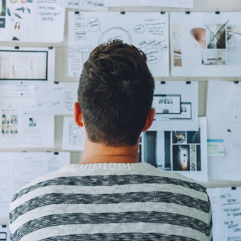 Image of man facing away from camera at a wall of various graphs, charts, and text pinned to the wall with thumbtacks.